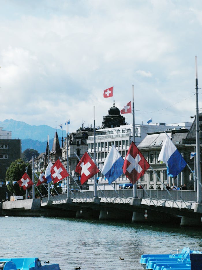 gallery-02 Picturesque view of Swiss flags on a bridge in Zurich with a backdrop of buildings and mountains.